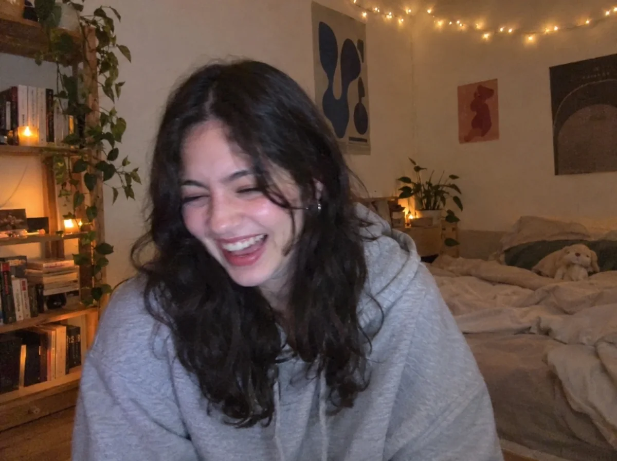 A woman with long dark wavy hair and a grey hoodie laughing with her eyes closed, in a bedroom with string lights, candles, plants and posters